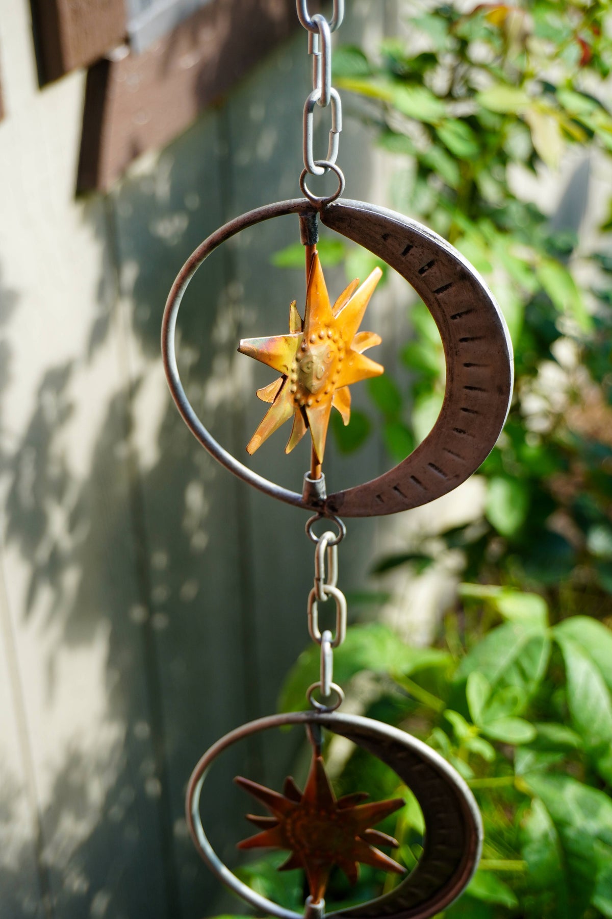 A metal garden rain chain with two interlocking crescent moons and starburst designs, featuring a bright orange flower in the center, hanging outdoors against a blurred background of foliage and a wall.
