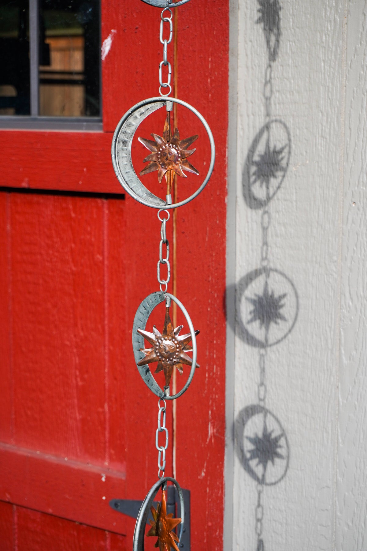 A hanging metal chain ornament with circular compass-style rings and starburst centers, shown outdoors against a red painted door and white wall.