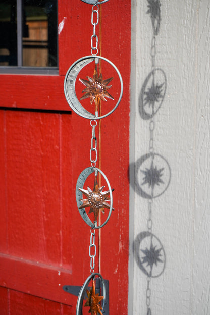 A hanging metal chain ornament with circular compass-style rings and starburst centers, shown outdoors against a red painted door and white wall.