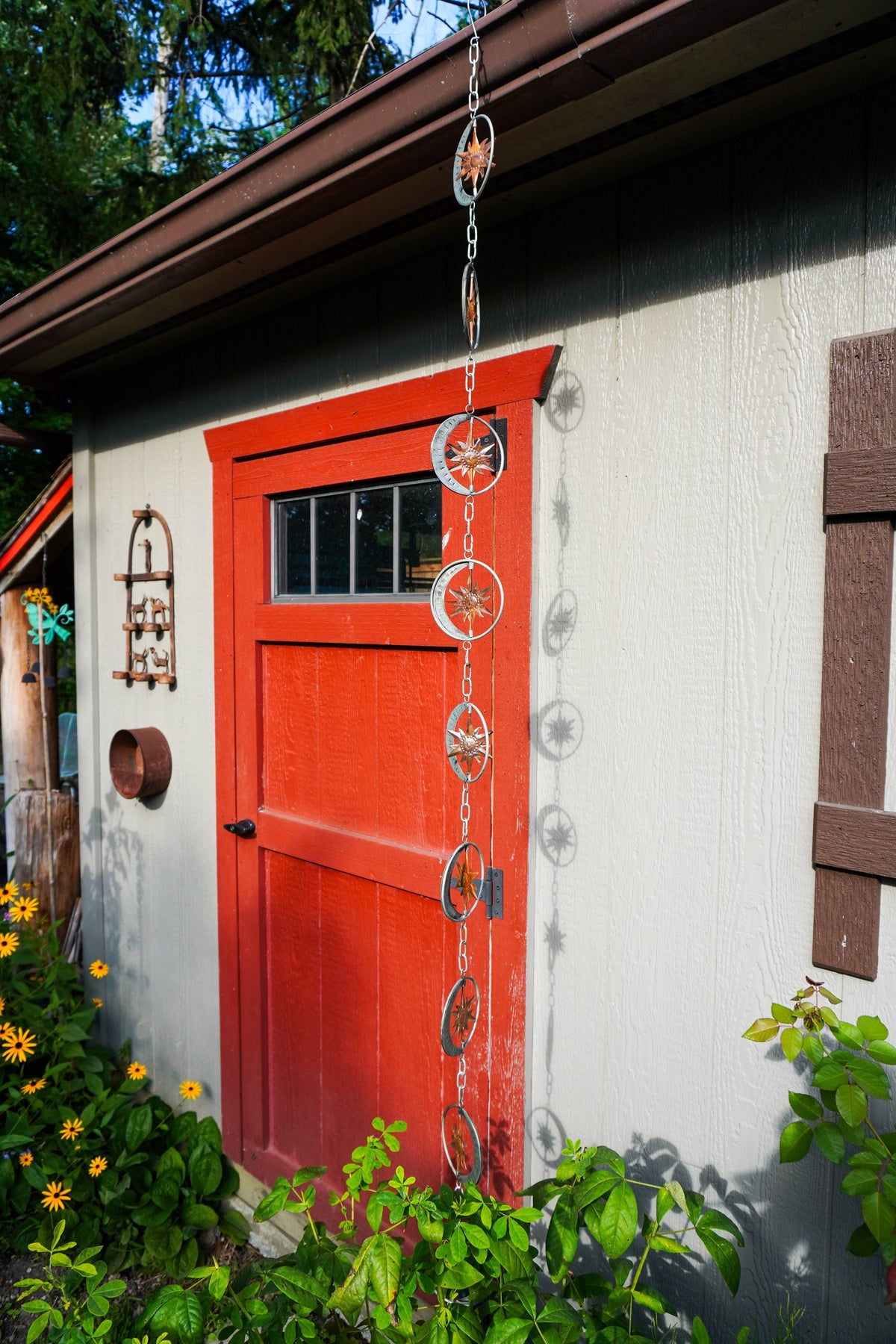 A decorative metal chain with circular compass-style discs hangs in front of a small white shed with a bright red door. Green plants and yellow flowers are visible at the base, and trees appear above the roofline.