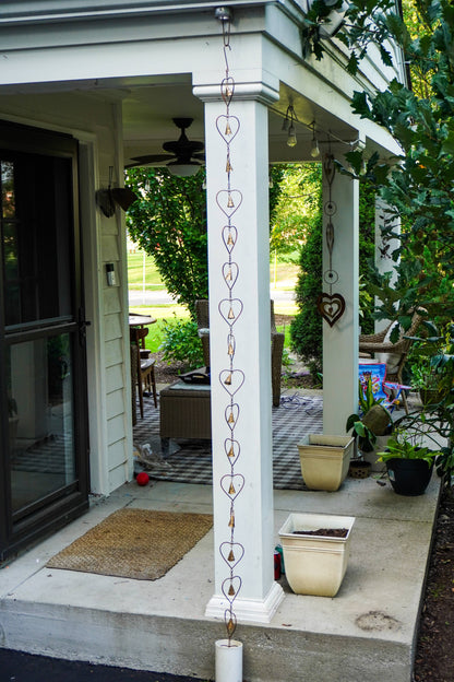 A decorative hanging heart-shaped metal wind chime hangs from a white porch column in a backyard setting.