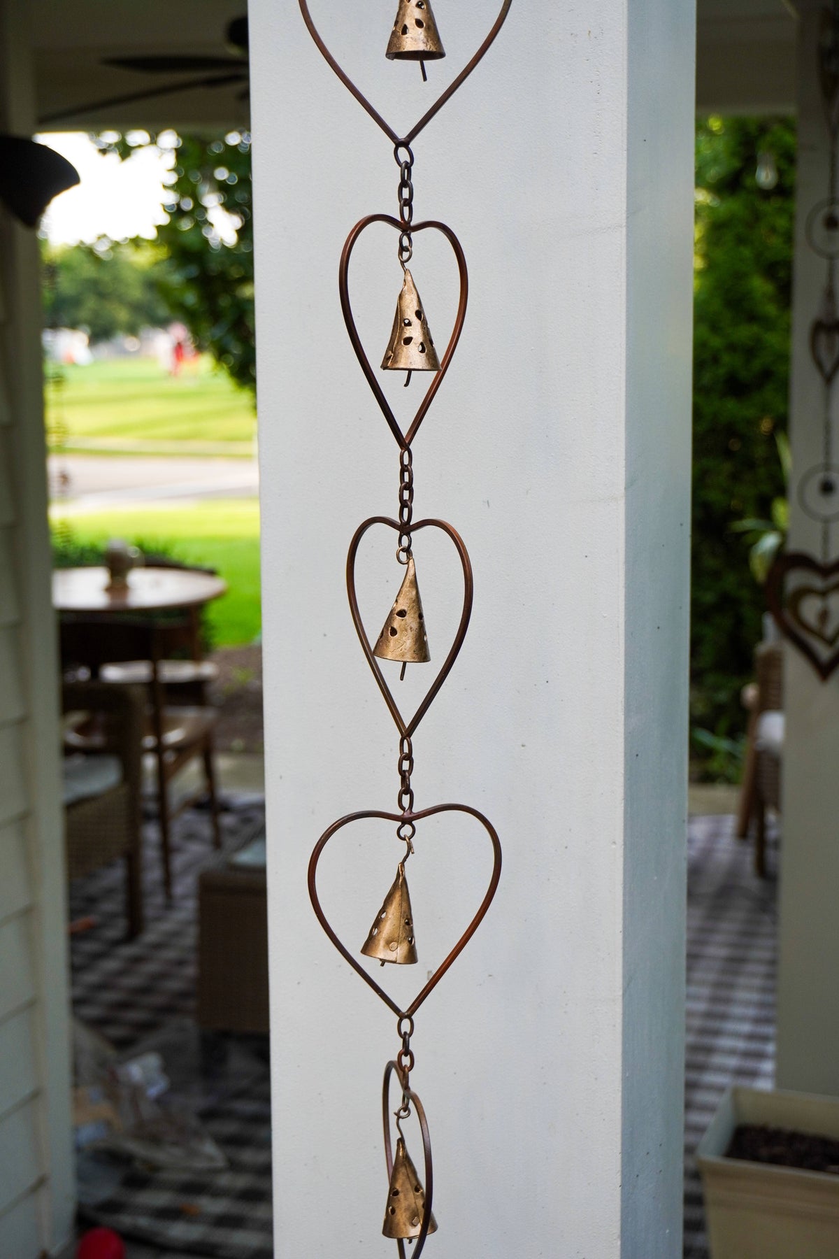 A rustic metal heart-shaped wind spinner hanging vertically on a white porch column, with a blurred outdoor garden and patio background.