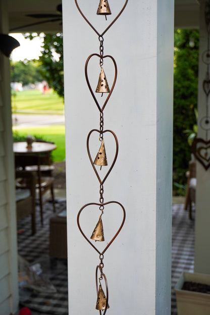 A rustic metal heart-shaped wind spinner hanging vertically on a white porch column, with a blurred outdoor garden and patio background.