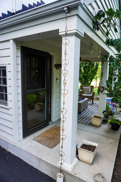 A decorative heart-shaped metal rain chain hanging vertically from a white porch column of a house. The porch has a black-framed glass door, a concrete walkway, potted plants, and outdoor seating visible in the background.