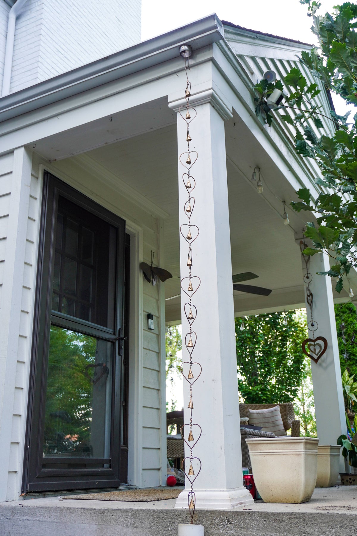 A decorative metal rain heart rain chain with interlocking heart shapes hangs from the eaves of a white porch with columns and a dark-framed front door.