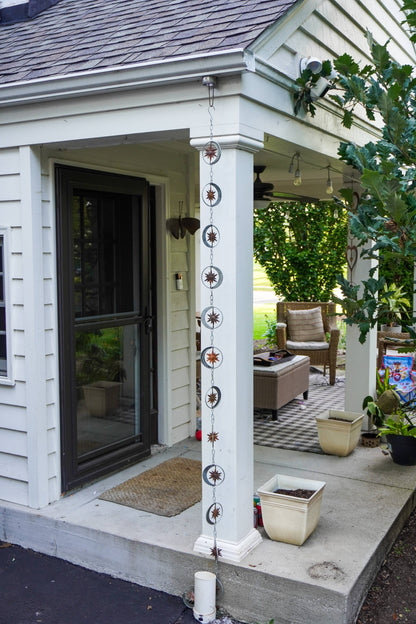 A decorative hanging chain of circular metal ornaments with starburst and floral motifs hangs vertically from a white porch post. The photo shows the exterior of a house porch with a sliding glass door, concrete steps, potted plants, and patio seating in the background.