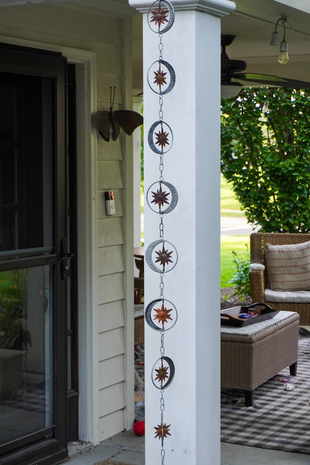 A vertical metal garden scroll hanging from a white porch column, featuring a repeating design of sunburst stars and crescent moons in silver and bronze tones, set against a residential porch background with a patio chair and greenery.