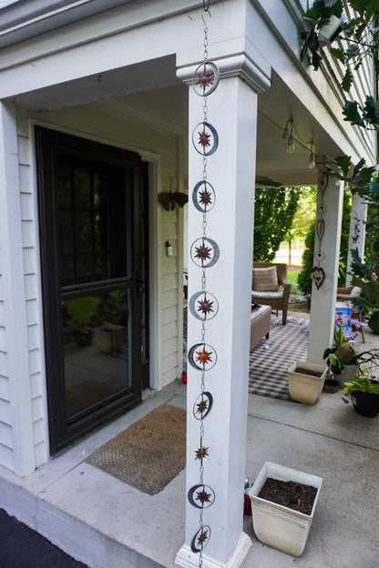 A decorative hanging chain with sun and moon metal medallions and starburst motifs displayed on a white porch column in front of a house entryway.