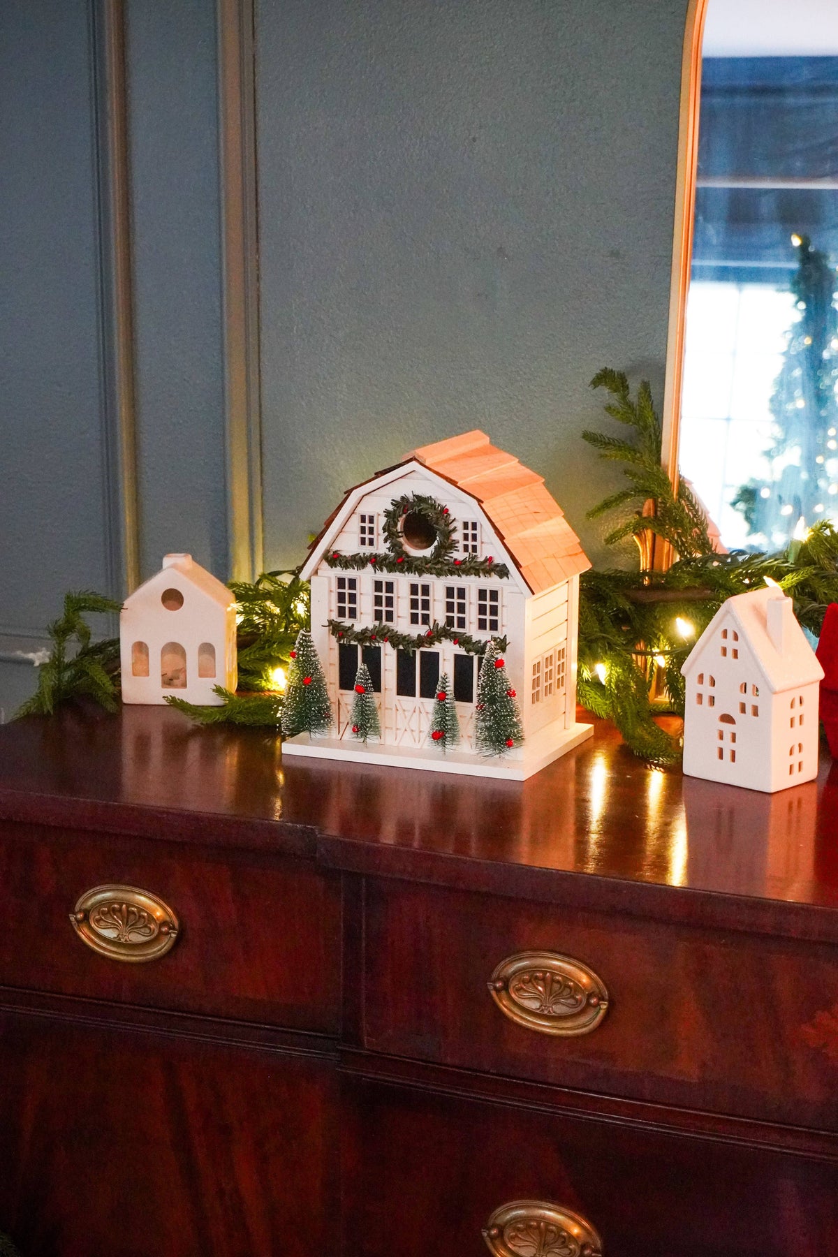A decorative white Christmas cottage ornament with a peach roof and green garland, displayed on a dark wood dresser alongside smaller matching ornaments and holiday greenery.