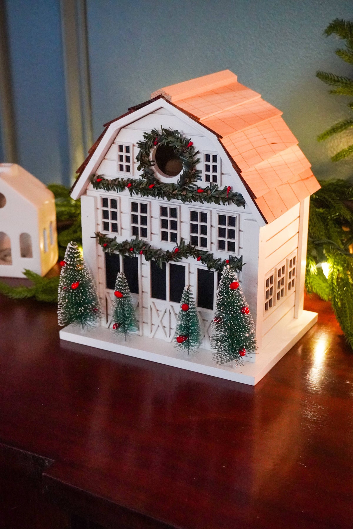 A white Christmas cottage ornament with a peach-colored roof, decorated with a green wreath and red berries, displayed on a dark wooden table with holiday lights in the background.