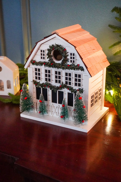 A white Christmas cottage ornament with a peach-colored roof, decorated with a green wreath and red berries, displayed on a dark wooden table with holiday lights in the background.