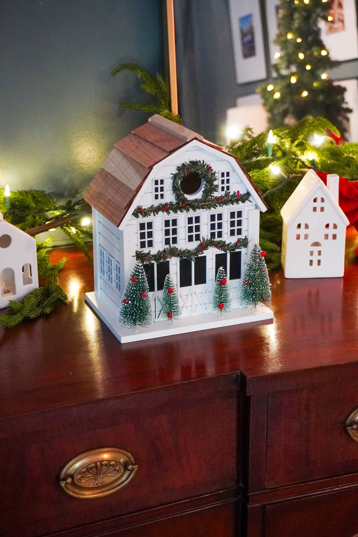 A decorative white farmhouse-style Christmas house ornament with a brown roof, black shutters, and small green trees with red ornaments, displayed on a dark wooden dresser with holiday lights and greenery in the background.