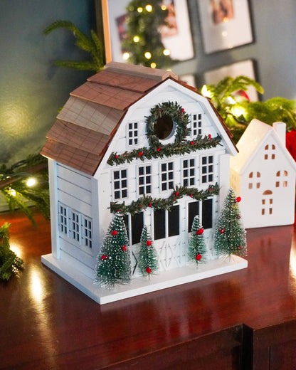 A decorative white house-shaped Christmas ornament or candle holder with a brown shingled roof, green garland, wreath, and small green Christmas trees with red ornaments, displayed on a wooden surface in a holiday setting.