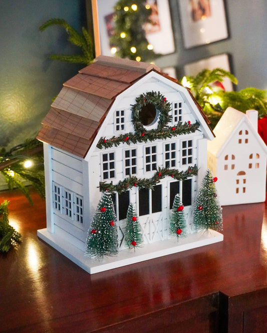 A decorative white house-shaped Christmas ornament or candle holder with a brown shingled roof, green garland, wreath, and small green Christmas trees with red ornaments, displayed on a wooden surface in a holiday setting.