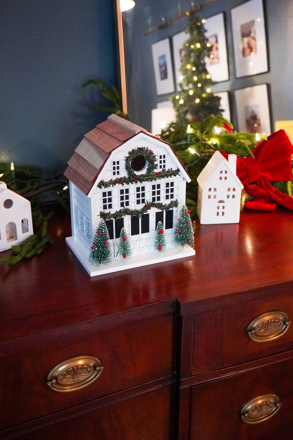 A white holiday cottage Christmas ornament with a brown roof and green garland details, displayed on a dark wood dresser alongside other festive decorations.
