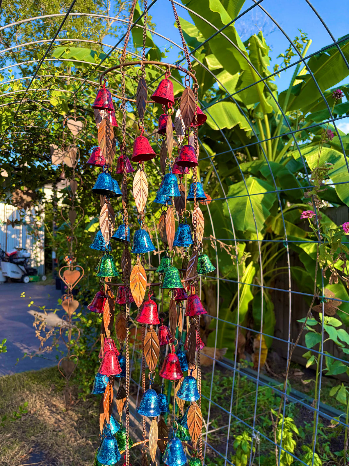 A colorful metal wind chime garden hanging ornament with red, blue, green, and gold bells and leaf-shaped charms, suspended in front of a wire trellis in a garden setting.