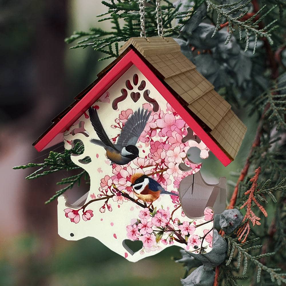 A decorative hanging birdhouse ornament with a pink cherry blossom design, two birds printed on the front, and a brown shingled roof, shown hanging among evergreen branches.