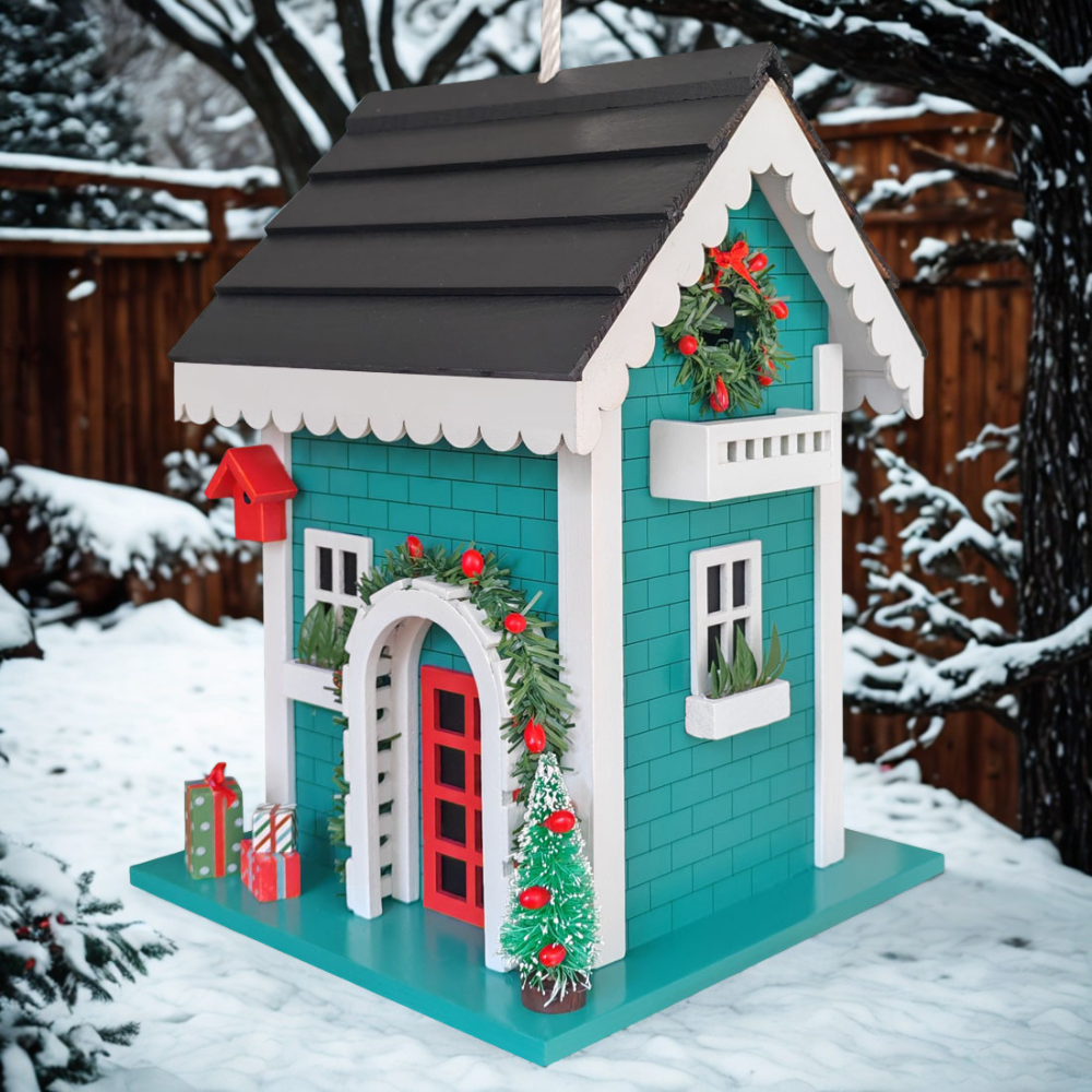 A decorative Christmas birdhouse or miniature house with teal siding, white trim, a dark gray roof, red door and accents, green wreaths, and a small tree on a snowy outdoor background.