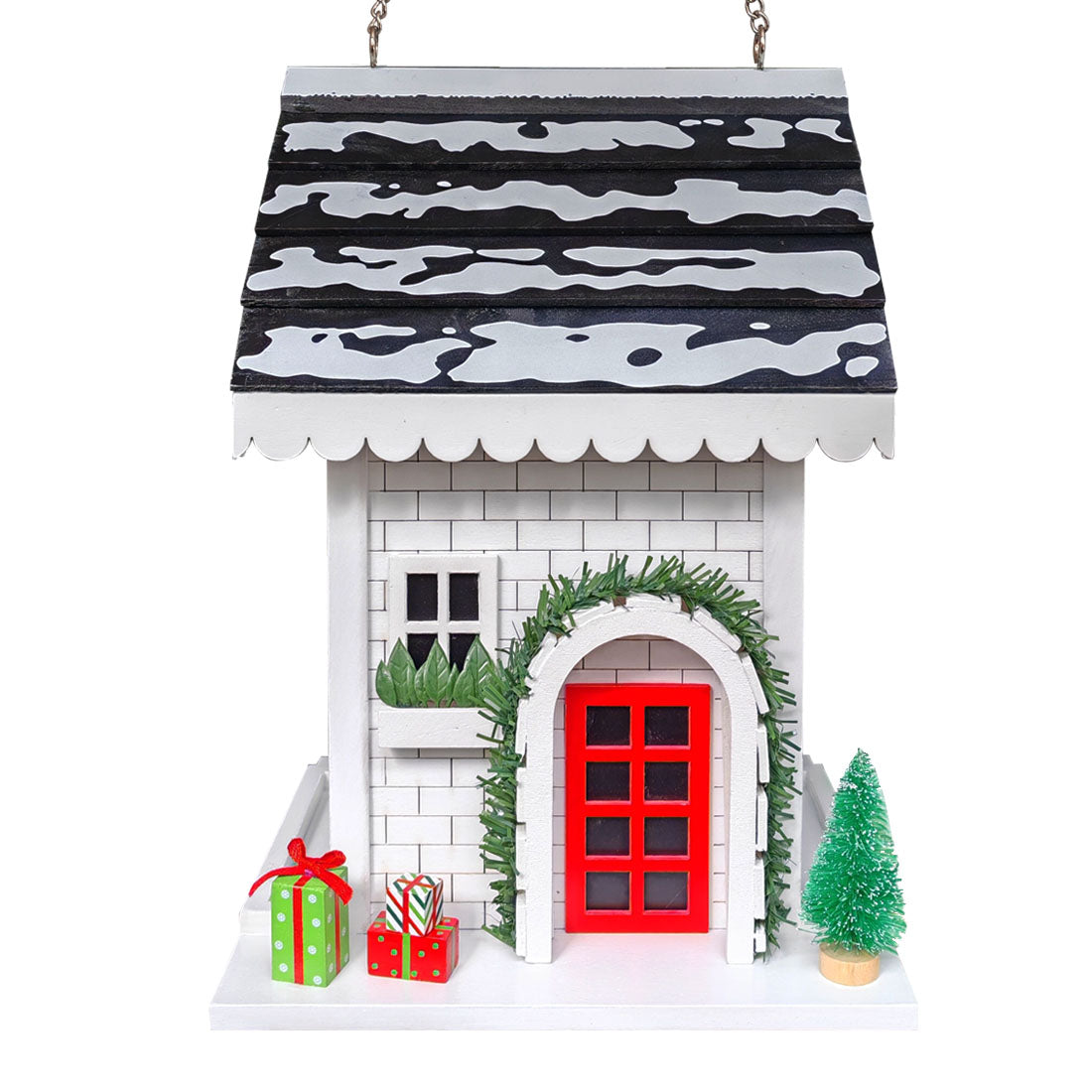 A decorative hanging Christmas bird feeder featuring a white brick facade, a black and white patterned roof, a bright red door, green garland around the doorway, small green gift boxes, and a miniature green tree on a white background.