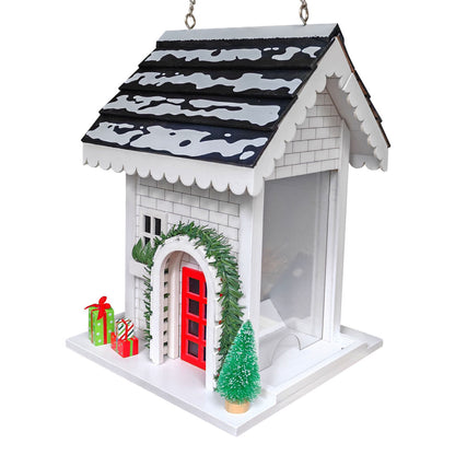 A white Christmas bird feeder shaped like a house, featuring a black and white patterned roof, a red front door, green garland, a small green tree, and holiday gifts on the side.
