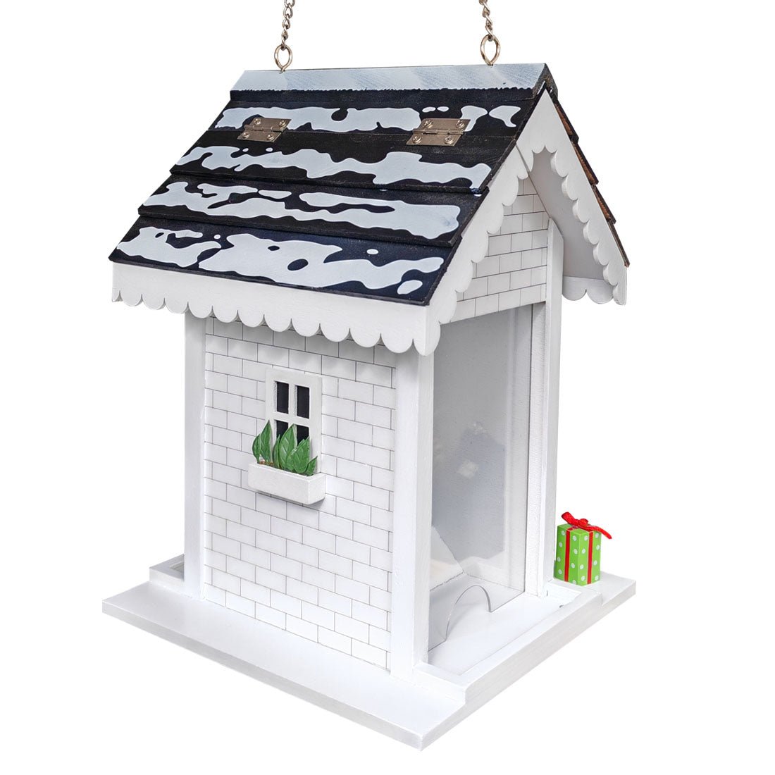 A white Christmas bird feeder shaped like a cottage, with a black and white snow-camouflage patterned roof, a small green plant in a window box, and a miniature green gift box with a red bow on the base.