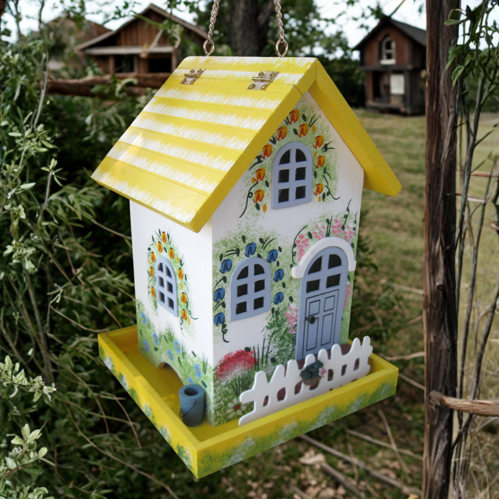 A decorative bird feeder shaped like a small house, hanging outdoors. The house has a yellow and white striped roof, a white body with painted floral and garden designs, a blue-gray front door, and a yellow tray at the bottom.