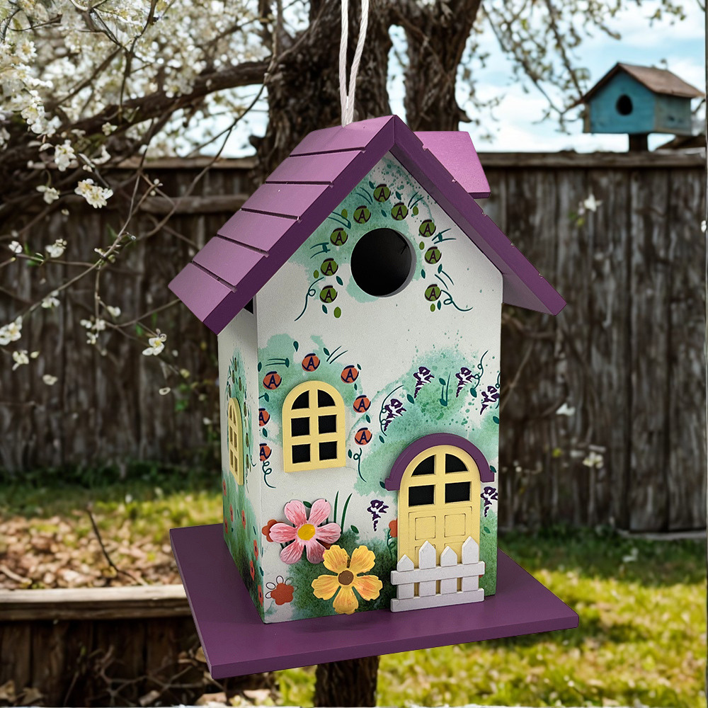 A decorative birdhouse shaped like a house, hanging outdoors in a garden. It has a purple roof and base, a white front, yellow window and door details, painted ladybugs, flowers, and a small white picket fence on the front.