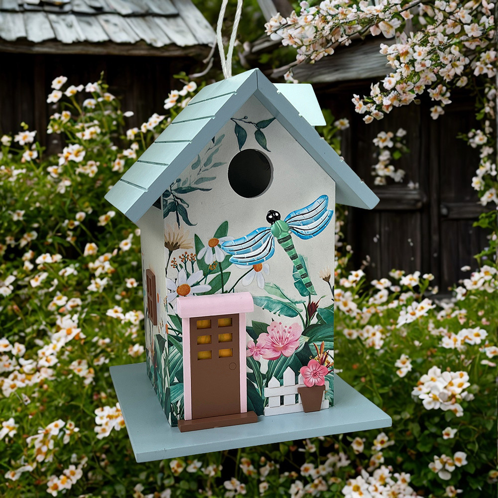A decorative birdhouse with a light blue roof and perch, decorated with a hand-painted tropical dragonfly and floral design. It features a small pink-framed brown door and is shown hanging in front of a garden with white flowers.
