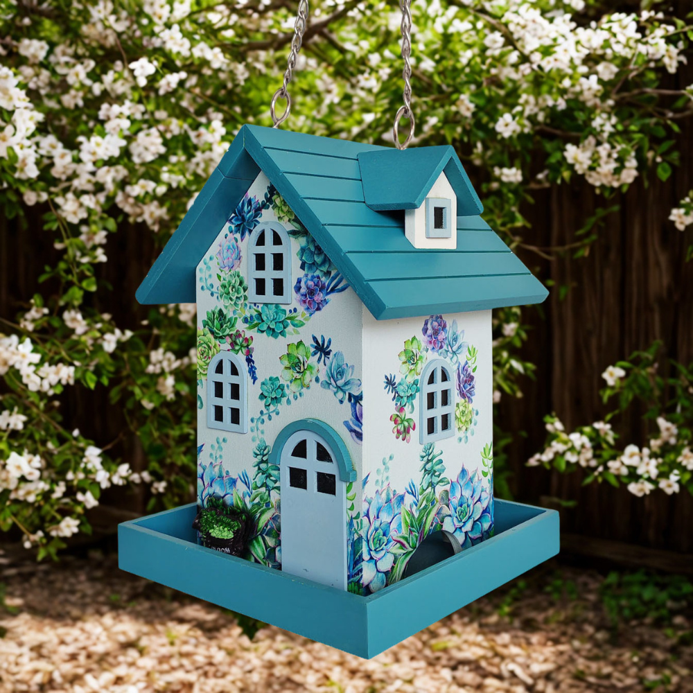 A decorative bird feeder shaped like a house, hanging from chains outdoors. The birdhouse has a teal roof and tray, a white body, and a floral print with blue, purple, and green flowers and leaves.