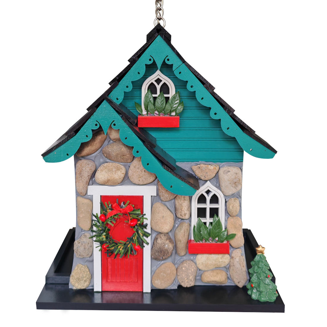A decorative Christmas bird house featuring a stone-textured gray facade, teal blue roof trim, a bright red front door with a green wreath, white arched windows, and small red planters with greenery, shown on a plain white background.
