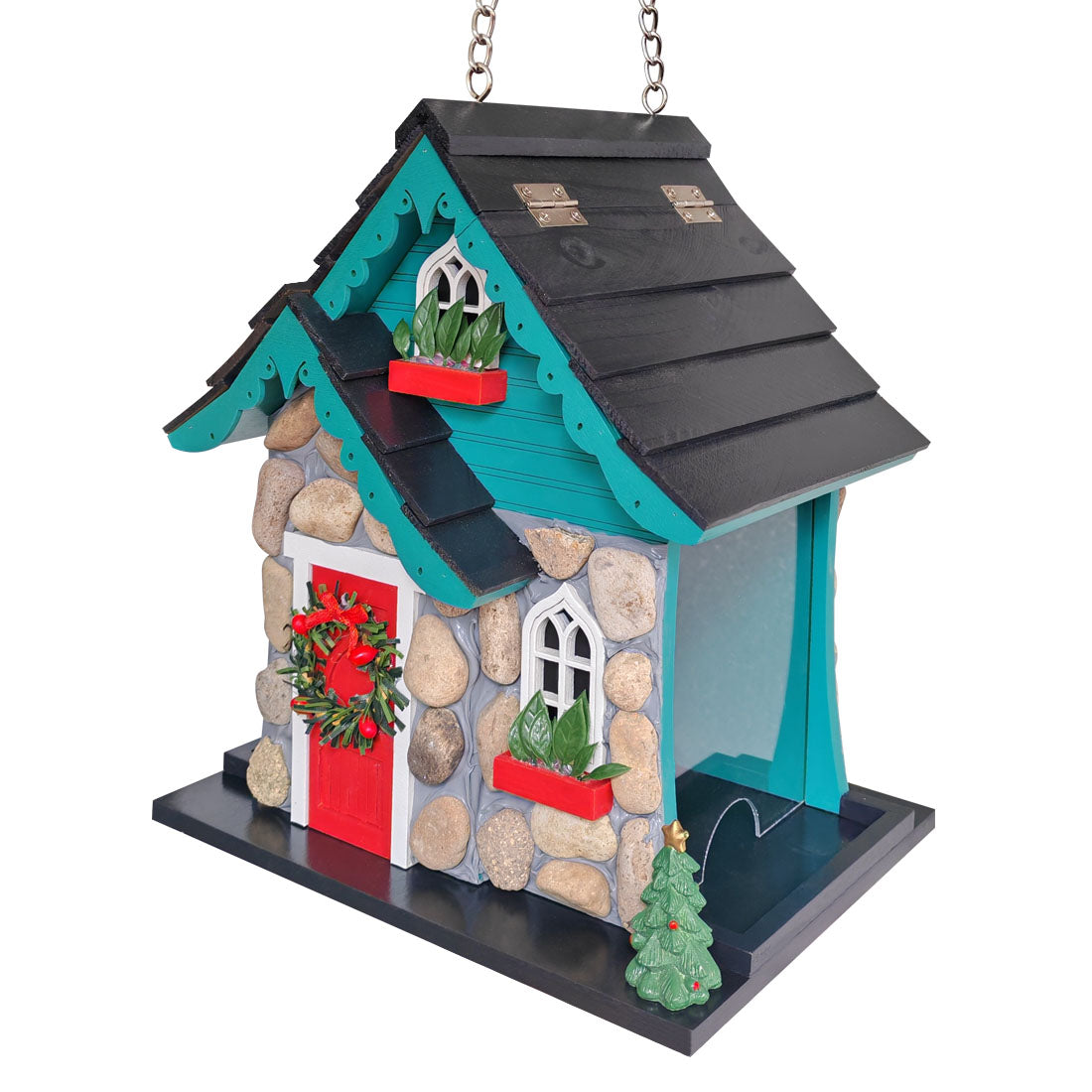 A decorative birdhouse shaped like a small house with a black shingled roof, teal trim, a stone-textured facade, a red door, white window frames, red planters with greenery, a red wreath, and a small green tree on a black base.