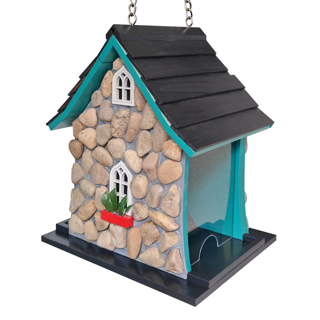 A decorative bird feeder shaped like a small house with a black shingled roof, faux-stone beige walls, turquoise trim, two white arched windows, a small red planter with green leaves, and a black feeding platform.