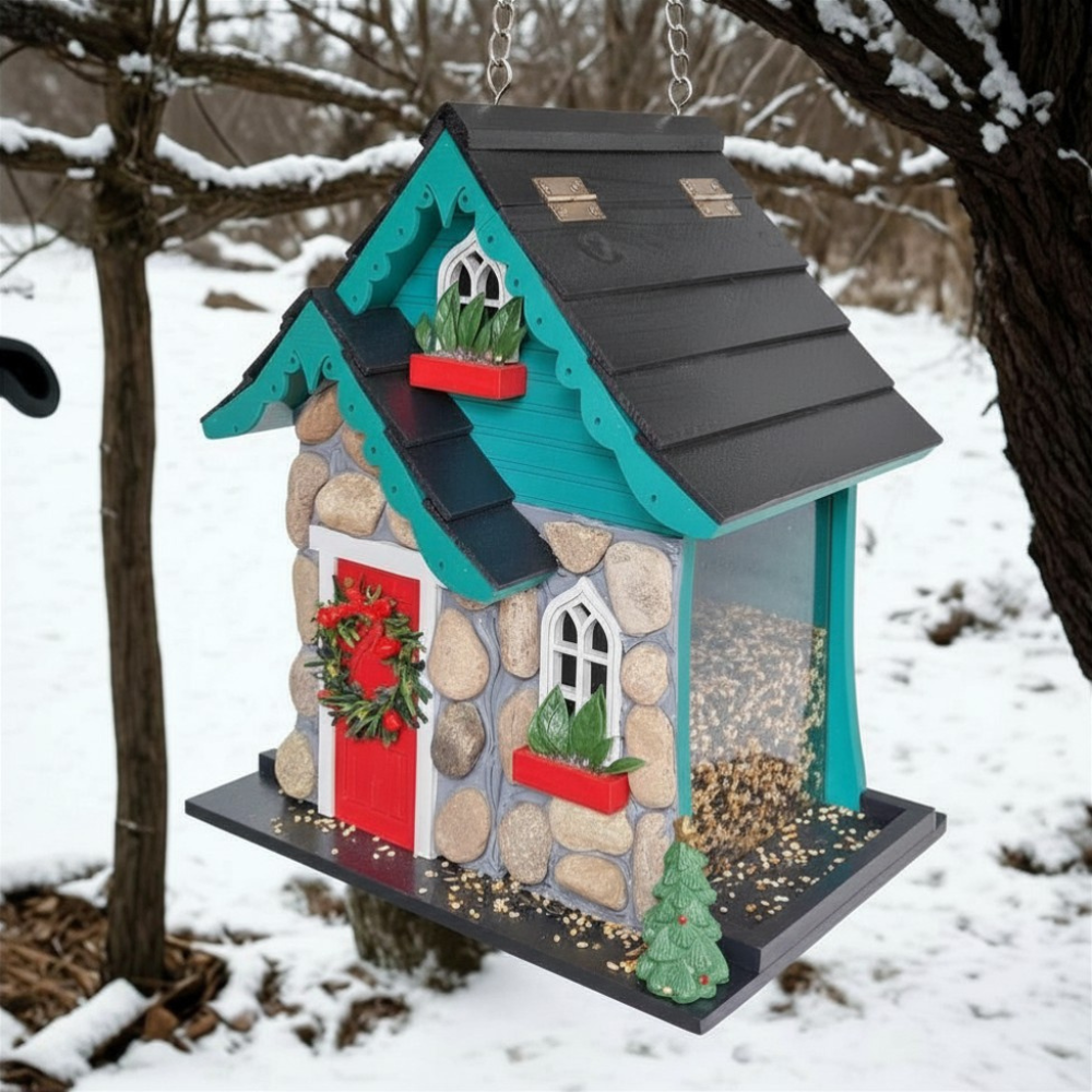 A decorative birdhouse shaped like a small house with a gray roof, faux-stone exterior, turquoise trim, a red door with a green wreath, and a seed hopper window, hanging in a snowy outdoor setting.