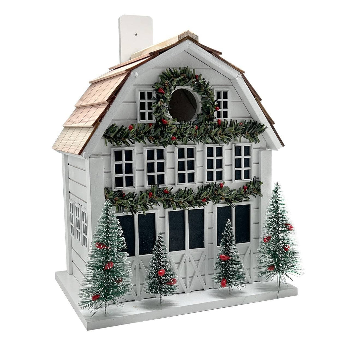 A white holiday barn birdhouse decorated with green garlands, red berries, small green Christmas trees, and a brown shingled roof, shown against a plain white background.