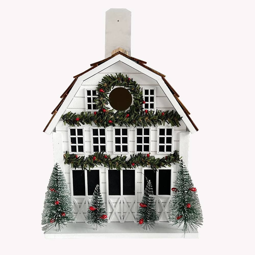 A white holiday house birdhouse decoration with a brown shingled roof, green garlands with red berries, a circular wreath on the front, and three small snow-covered Christmas trees at the base, shown on a plain white background.