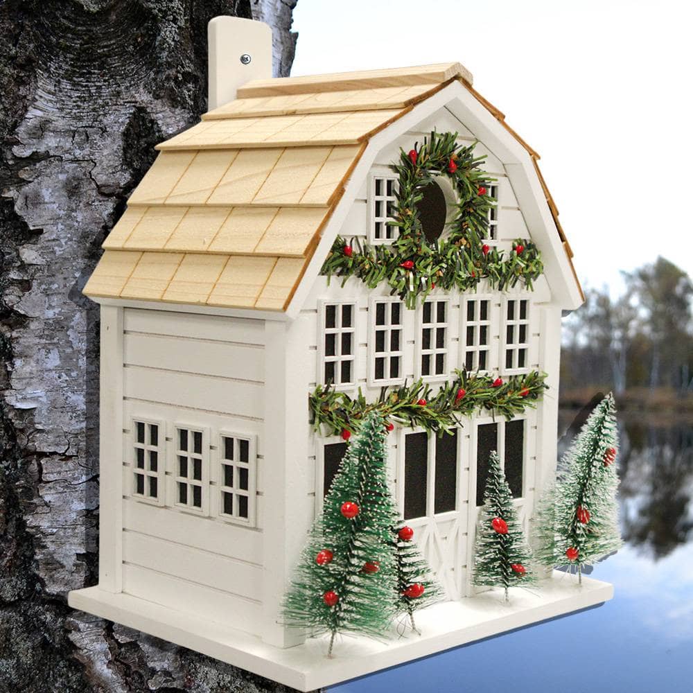 A decorative white Christmas house birdhouse with a light wood shingled roof, green garlands with red berries, and three small frosted Christmas trees, mounted against a tree trunk outdoors.
