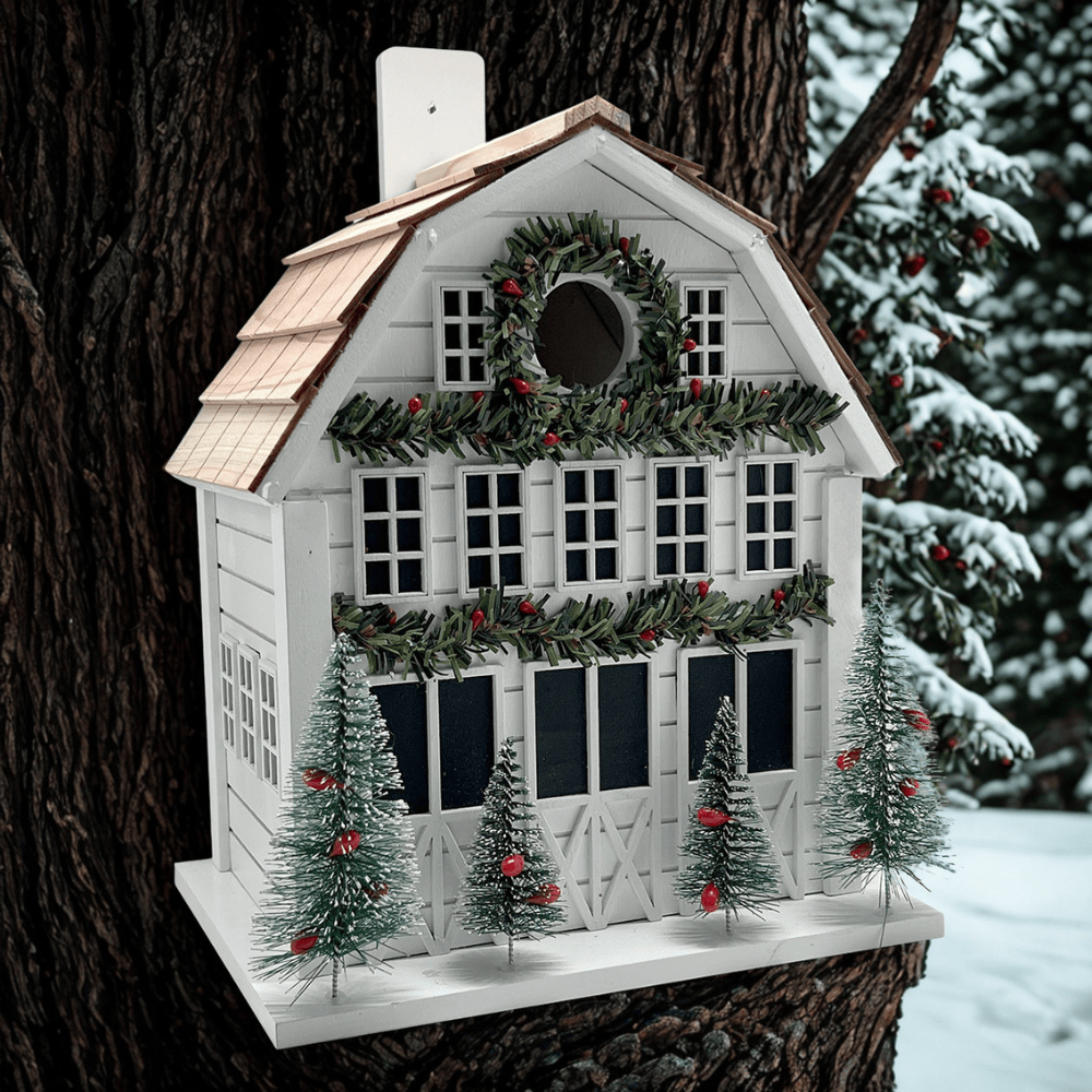 A white holiday barn birdhouse with a green garland wreath, red berries, and four small decorated Christmas trees, mounted on a tree trunk in a snowy outdoor setting.