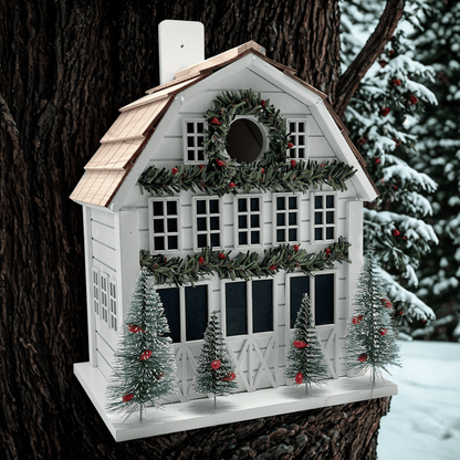 A white holiday barn birdhouse with a green garland wreath, red berries, and four small decorated Christmas trees, mounted on a tree trunk in a snowy outdoor setting.