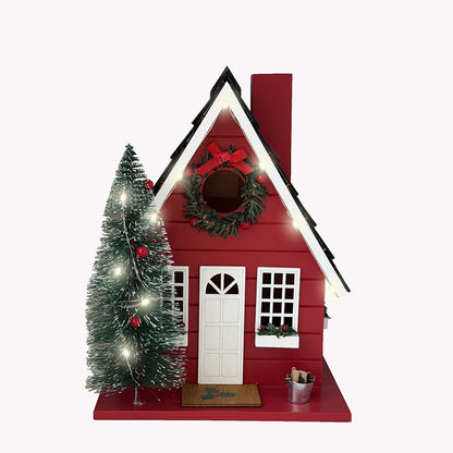 A red Christmas bird house figurine with a white door, white-framed windows, a green wreath with a red bow on the front, a small decorated Christmas tree beside it, and warm white string lights, shown on a plain light background.