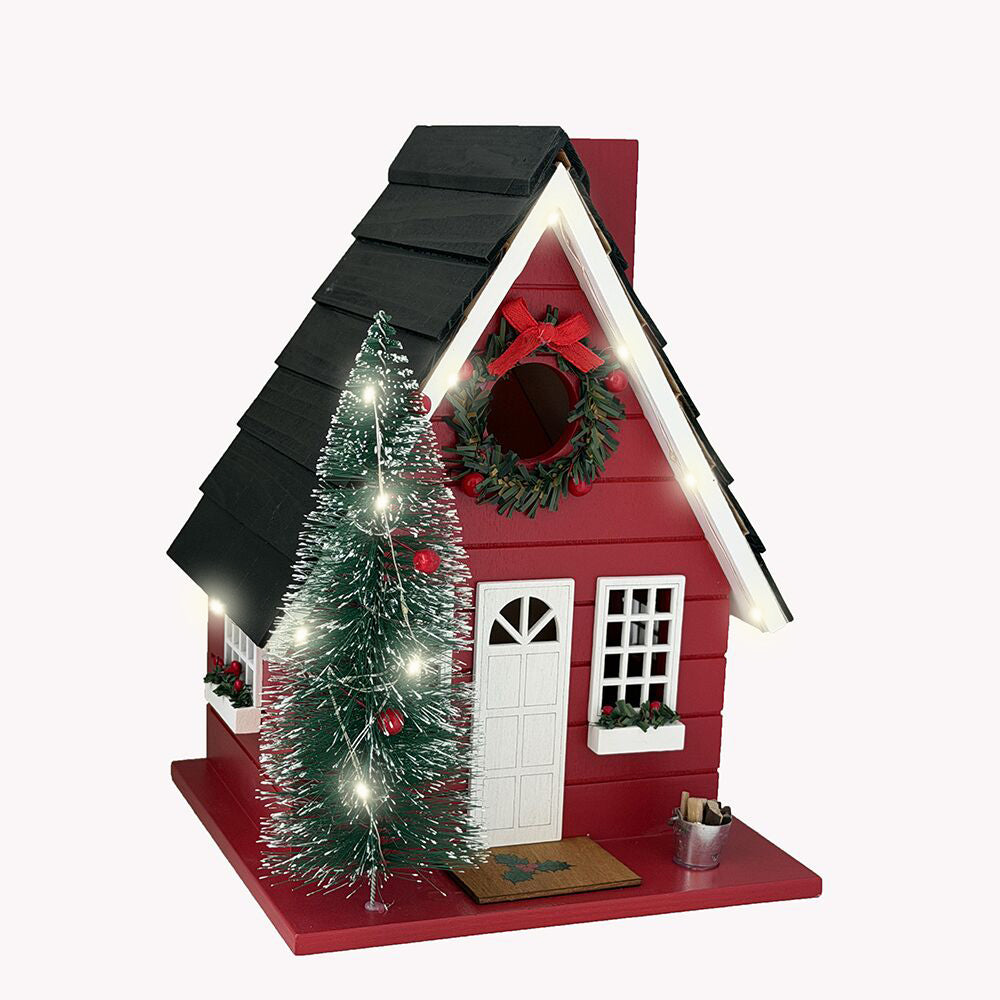 A red Christmas birdhouse with a black shingled roof, white door and windows, a green wreath with a red bow on the front, and a small artificial Christmas tree with white lights and red ornaments on the porch.