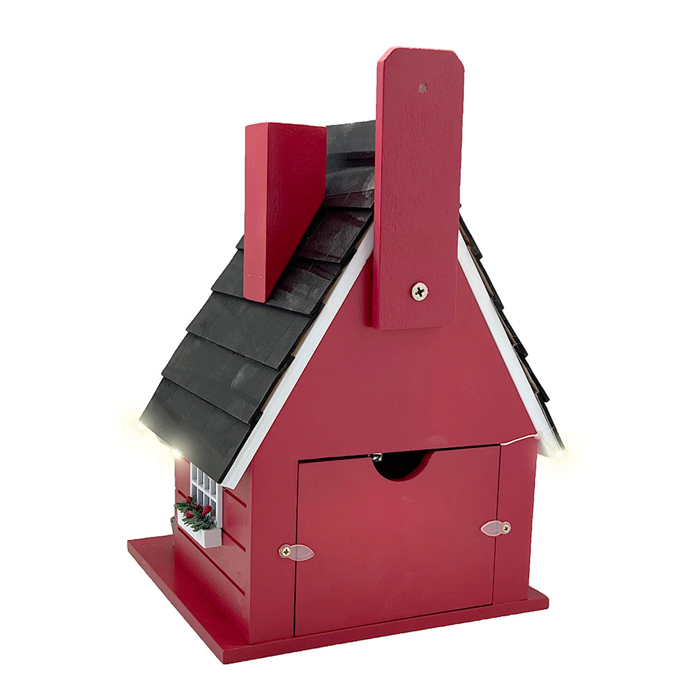 A red birdhouse shaped like a cottage with a black shingled roof, small front door, and a small light glowing from the side, shown on a plain white background.