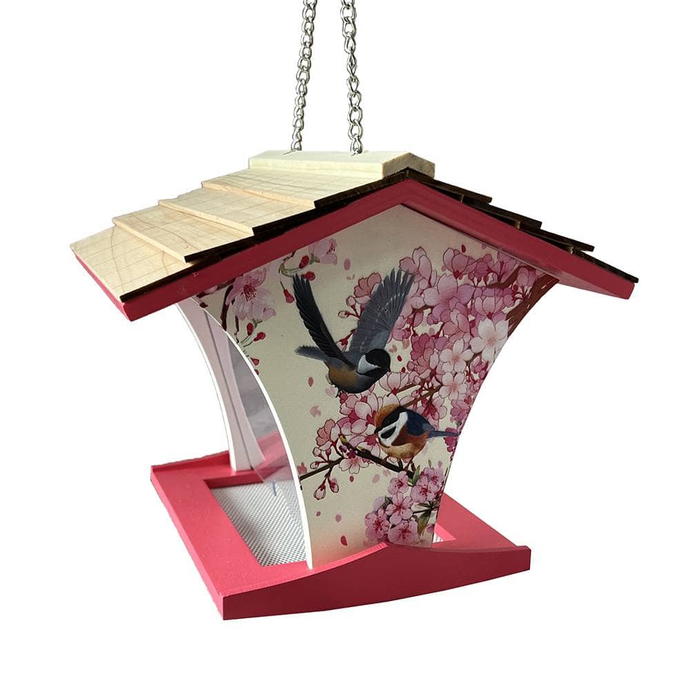 A hanging bird feeder with a pink and cherry blossom printed panel, featuring two birds in flight and perched on branches. The feeder has a light wood slatted roof, a bright pink base, and is suspended by a silver metal chain against a plain white background.