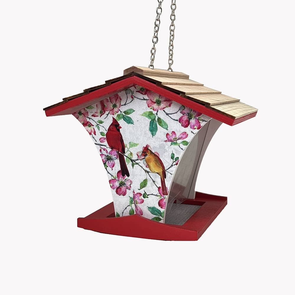 A hanging bird feeder with a red roof and base, a light wood shingled top, and a white front panel printed with pink blossoms and two birds, suspended by a metal chain against a plain white background.