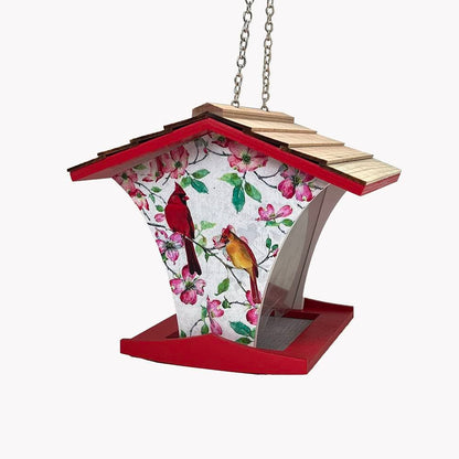 A hanging bird feeder with a red roof and base, a light wood shingled top, and a white front panel printed with pink blossoms and two birds, suspended by a metal chain against a plain white background.