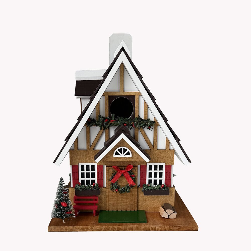 A decorative miniature Christmas bird house with a steep gabled roof, white and wood trim, red shutters, a red bow on the door, green garlands, a small tree, bench, and log props on a plain light background.