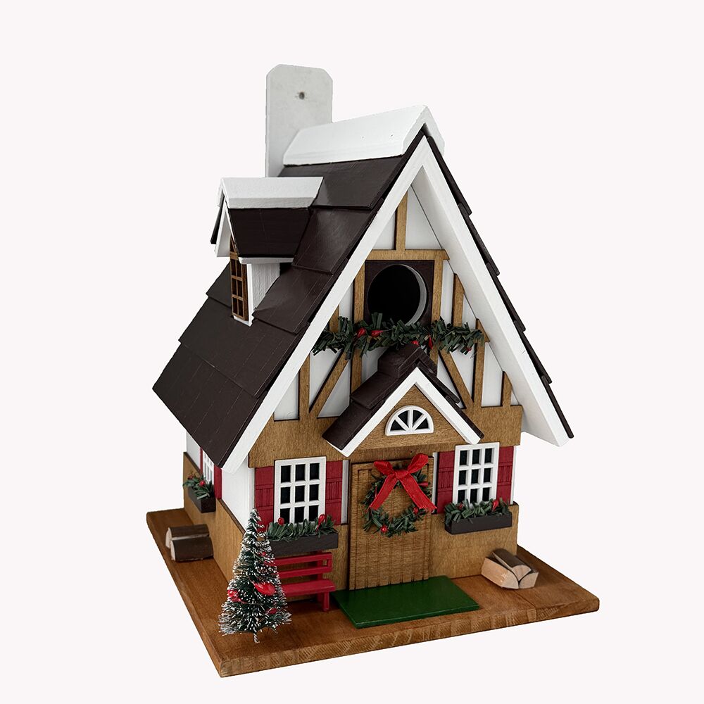 A miniature Christmas gingerbread bird house ornament or figurine with a dark brown roof, white and wood trim, red shutters, a green wreath with a red bow, a small Christmas tree, and a bench on a wooden base, shown against a plain light background.