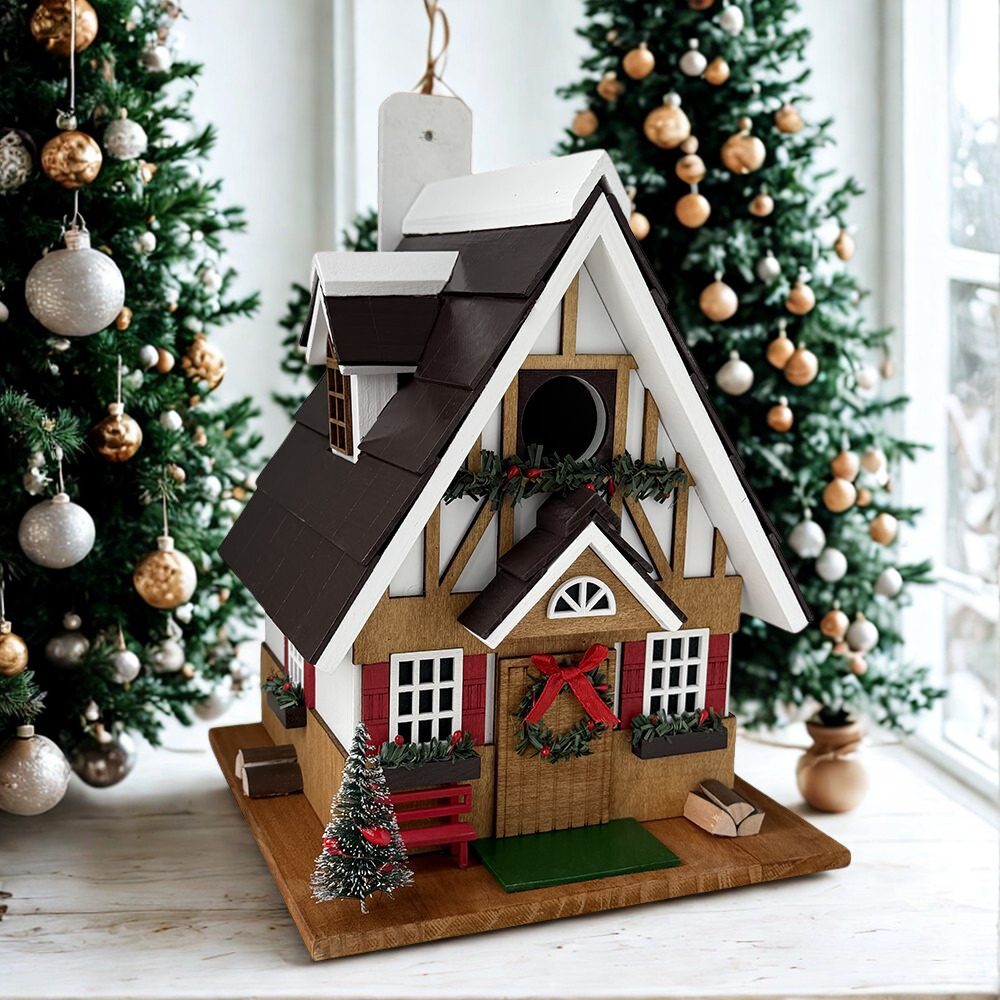 A decorative gingerbread birdhouse styled as a Christmas cottage with a dark brown roof, white trim, red shutters, a wooden door with a red bow, and a small tree on a base, set in front of decorated Christmas trees.