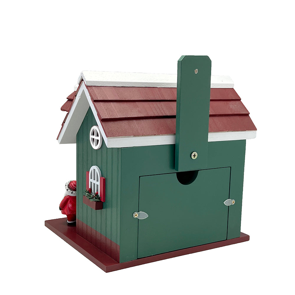 A Christmas-themed wooden birdhouse shaped like a green cabin with a reddish-brown shingled roof, white trim, silver metal hinges, and a small Santa figure standing on the left side.