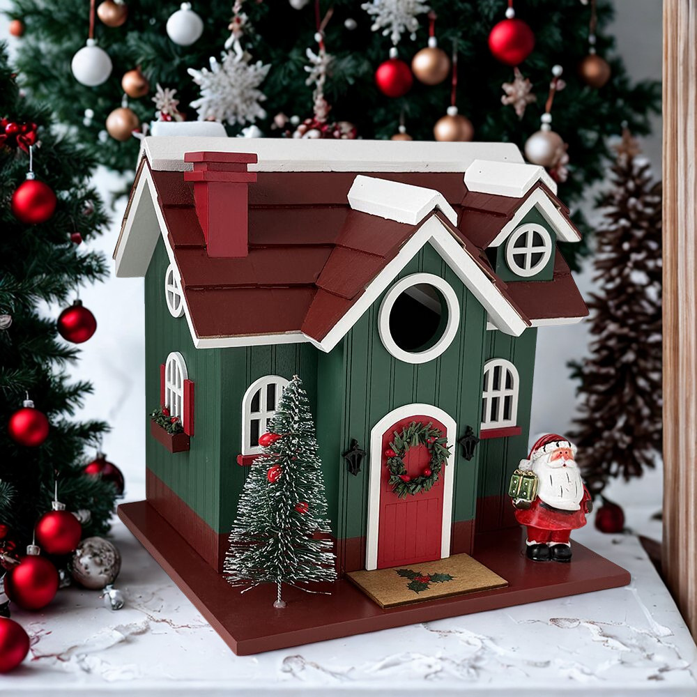 A decorative wooden birdhouse with a green body, red roof, white trim, a small Christmas tree, wreath, and Santa figure, displayed in front of a decorated Christmas tree.