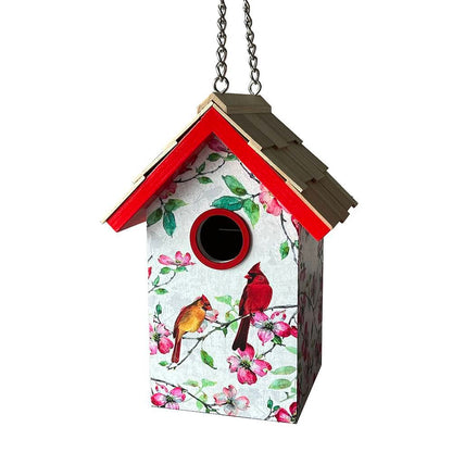 A decorative hanging birdhouse with a white floral front panel featuring two cardinals and pink blossoms, a red-trimmed gabled roof, a brown shingled top, and a metal hanging chain against a plain white background.
